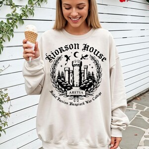 Puede incluir: Sudadera color crema con un gr&aacute;fico negro de un castillo, &aacute;rboles y el texto "Riorkson House Aretia Rebel Faction Basgiath War College". El dise&ntilde;o est&aacute; rodeado por una corona de laurel.