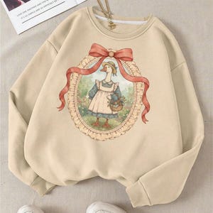 May include: Beige sweatshirt featuring a whimsical illustration of a goose wearing a bonnet and apron, holding a basket of flowers. The artwork is framed by a red ribbon and an oval border. The sweatshirt has long sleeves and a crew neck.