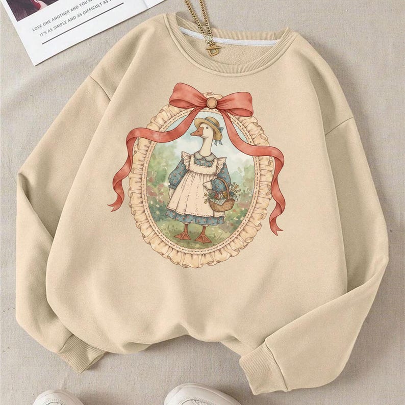 May include: Beige sweatshirt featuring a whimsical illustration of a goose wearing a bonnet and apron, holding a basket of flowers. The artwork is framed by a red ribbon and an oval border. The sweatshirt has long sleeves and a crew neck.