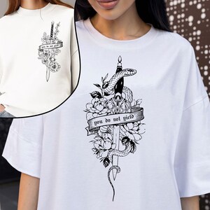May include: White t-shirt featuring a black and white graphic design. The design includes a sword, snake, flowers, and a banner with the text "you do not yield". The shirt has short sleeves and a crew neck.