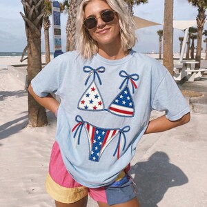 Peut inclure: T-shirt bleu clair orné d'un motif de bikini patriotique avec des étoiles et des rayures rouges, blanches et bleues. Le t-shirt est associé à un short coloré. Le motif rappelle le drapeau américain.