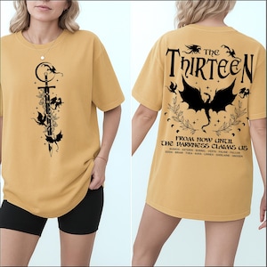 May include: Mustard yellow t-shirt with a black graphic on the front featuring a sword, flowers, and bats. The back of the shirt has the text "The Thirteen" above a dragon graphic and the phrase "From Now Until The Darkness Claims Us."