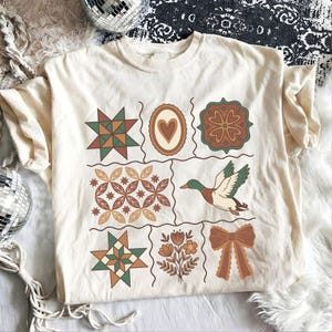 以下が含まれることがあります： クリーム色のTシャツ。パッチワークのデザインで、緑、茶色、オレンジの色合いの幾何学模様と花柄が特徴です。飛んでいるアヒルと茶色のリボンも含まれています。