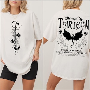 May include: White t-shirt featuring a black graphic of a sword, dragons, and floral elements on the front. The back of the shirt displays the text "The Thirteen" and "From now until the darkness claims us" with dragon and floral designs.