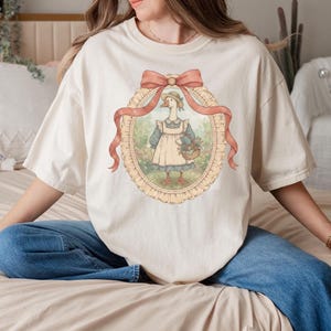 May include: Cream-colored t-shirt featuring a framed illustration of a goose wearing a bonnet and apron, carrying a basket of flowers. The illustration is framed with a peach-colored ribbon and a decorative oval border. The shirt has short sleeves and a relaxed fit.
