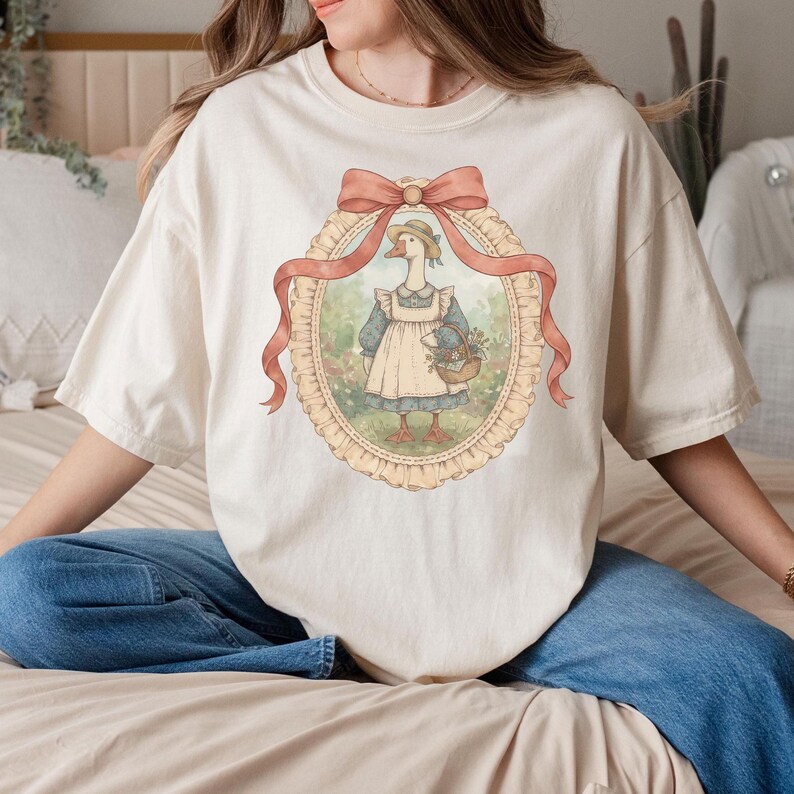 May include: Cream-colored t-shirt featuring a framed illustration of a goose wearing a bonnet and apron, carrying a basket of flowers. The illustration is framed with a peach-colored ribbon and a decorative oval border. The shirt has short sleeves and a relaxed fit.