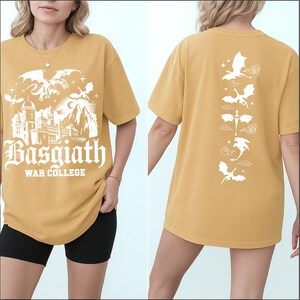 May include: Mustard yellow t-shirt with a white graphic on the front featuring a castle, dragons, and the text "Basgiath War College." The back of the shirt displays a vertical line of dragon illustrations. A casual, comfortable tee.