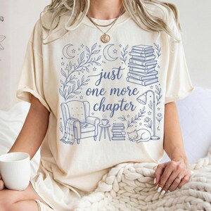 Könnte beinhalten: Cremefarbenes T-Shirt mit blauer Illustration und dem Text "just one more chapter". Das Design umfasst einen Bücherstapel, einen Sessel, eine Katze und florale Elemente. Ein tolles Geschenk für Leser.