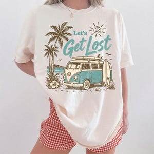 May include: An off-white short-sleeve t-shirt featuring a graphic print of a blue and white vintage van with a surfboard, palm trees, and the text Let's Get Lost in teal script. The design includes a sun icon and a beach scene in brown ink.