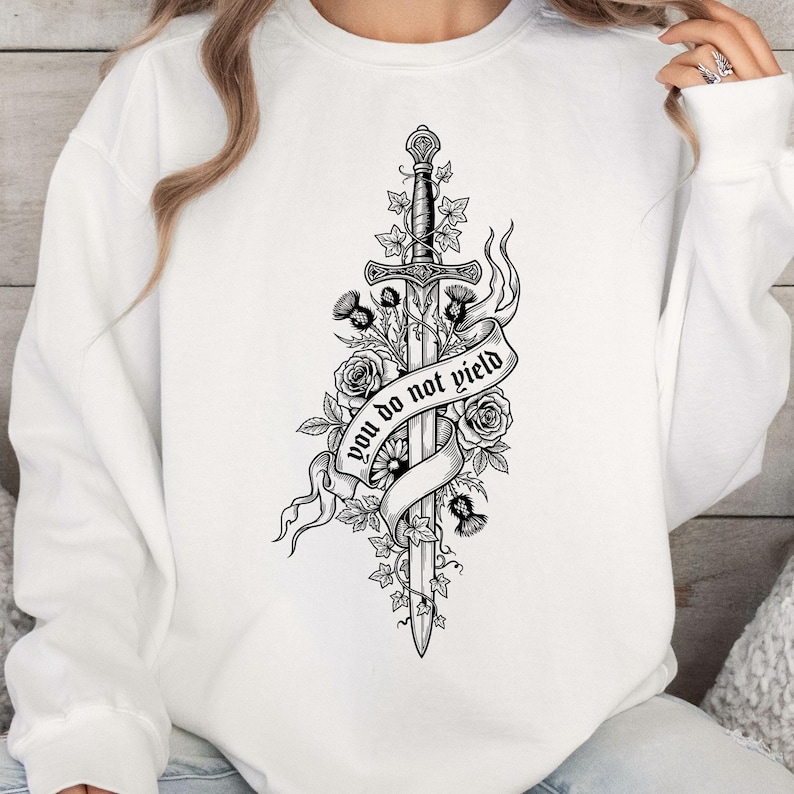May include: White sweatshirt featuring a black and white illustration of a sword entwined with roses, thistles, and ivy. A banner across the sword reads "you do not yield". The design is centered on the front of the garment.