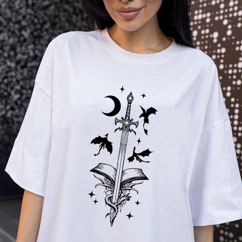 Puede incluir: Camiseta blanca con un gr&aacute;fico negro de una espada que atraviesa un libro abierto, con una luna creciente, murci&eacute;lagos y estrellas. El dise&ntilde;o tiene un tema de fantas&iacute;a.