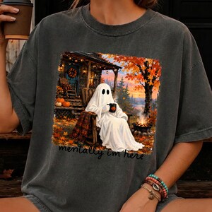May include: A charcoal gray t-shirt featuring a graphic of a white ghost sitting in a wooden chair by a campfire. The scene includes autumn trees, pumpkins, and a cabin. The text "mentally i'm here" is printed in black script below the graphic.