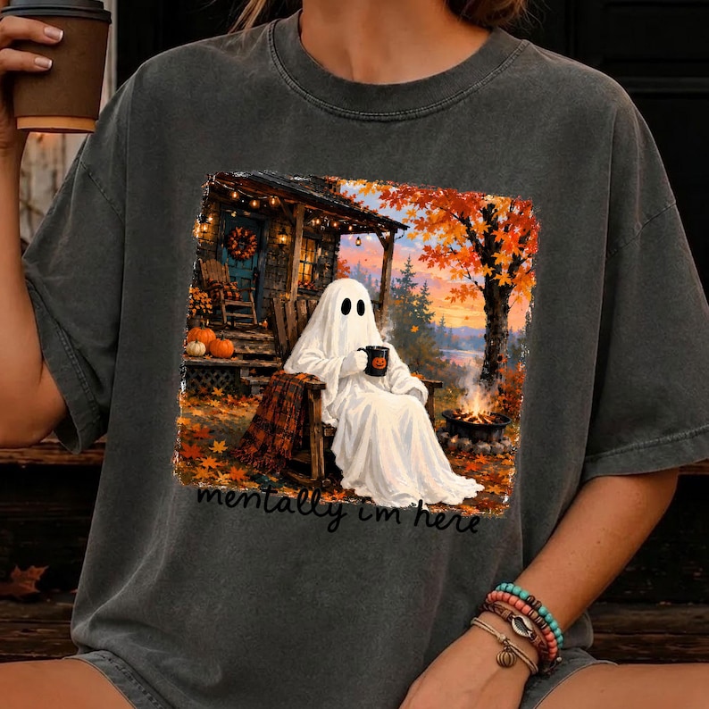 May include: A charcoal gray t-shirt featuring a graphic of a white ghost sitting in a wooden chair by a campfire. The scene includes autumn trees, pumpkins, and a cabin. The text "mentally i'm here" is printed in black script below the graphic.