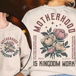May include: Cream-colored sweatshirt with the words "MOTHERHOOD" and "KINGDOM WORK" in a circular design. A floral illustration in pink and beige is centered below the text. The sweatshirt also features a Bible verse.