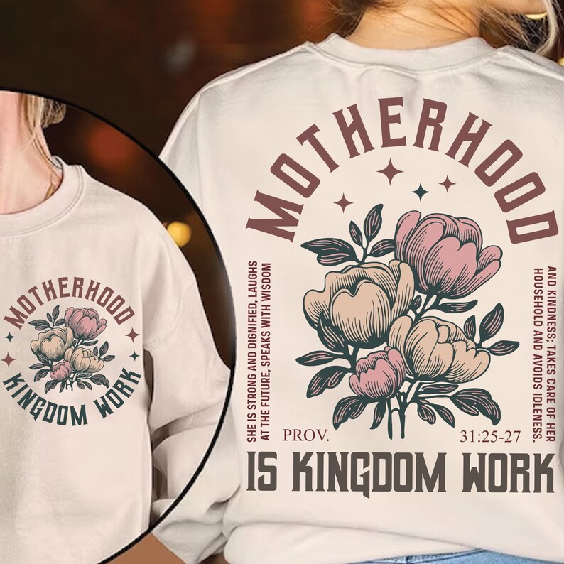 May include: Cream-colored sweatshirt with the words "MOTHERHOOD" and "KINGDOM WORK" in a circular design. A floral illustration in pink and beige is centered below the text. The sweatshirt also features a Bible verse.