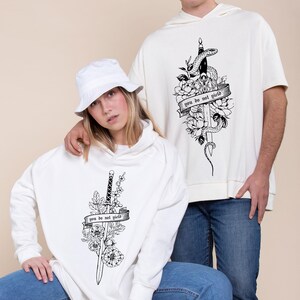 May include: White hooded sweatshirts with black graphic designs. One features a sword, flowers, and the text "you do not yield." The other has a snake, sword, flowers, and the same text. Both are worn by people.