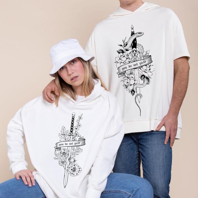 May include: White hooded sweatshirts with black graphic designs. One features a sword, flowers, and the text "you do not yield." The other has a snake, sword, flowers, and the same text. Both are worn by people.