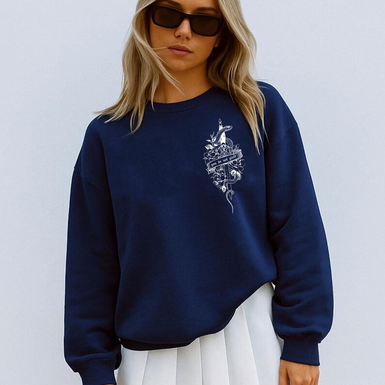 May include: Navy blue sweatshirt with a white graphic design on the upper left chest. The graphic includes a snake, flowers, and the text "we are not girls". The sweatshirt is paired with a white pleated skirt.