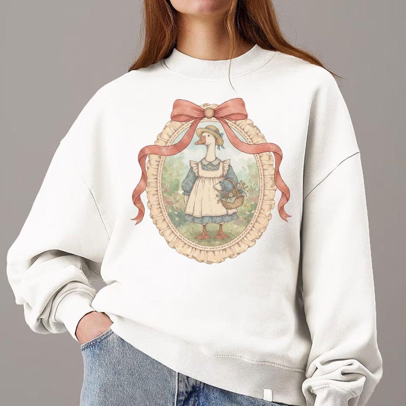 May include: White sweatshirt featuring a framed illustration of a goose wearing a bonnet and apron, holding a basket of flowers. The illustration is surrounded by a pink ribbon and a decorative oval frame. The sweatshirt has a crew neck and long sleeves.