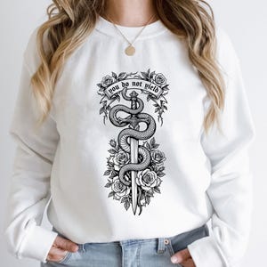 May include: White sweatshirt featuring a black and white graphic of a sword entwined by a snake, surrounded by roses. The text "you do not yield" is displayed above the sword. The design is centered on the front of the sweatshirt.