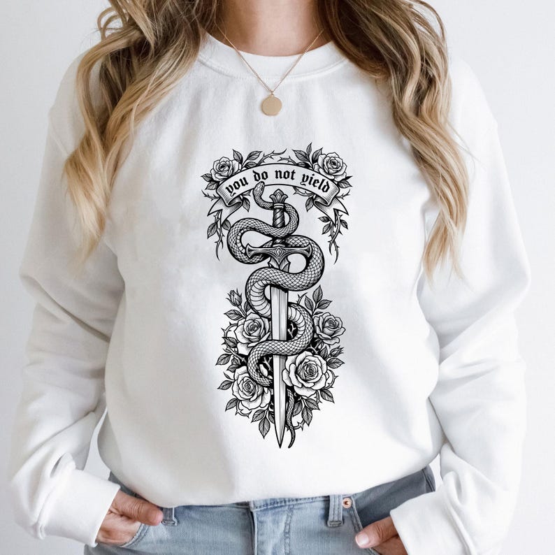 May include: White sweatshirt featuring a black and white graphic of a sword entwined by a snake, surrounded by roses. The text "you do not yield" is displayed above the sword. The design is centered on the front of the sweatshirt.