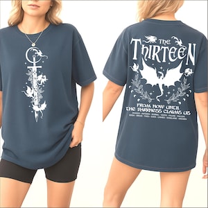 May include: Dark blue t-shirt with a white graphic on the front featuring a sword, flowers, and winged creatures. The back of the shirt displays the text "The Thirteen" above a winged creature design and the phrase "From Now Until The Darkness Claims Us."