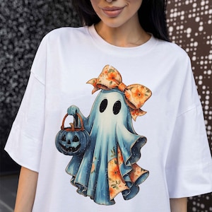 Op de afbeelding: Een wit t-shirt met een afbeelding van een lichtblauwe geest die een grote oranje bloemenstrik op het hoofd draagt. De geest houdt een donkerblauwe pompoenemmer vast en de onderkant van het laken van de geest toont een oranje bloemenpatroon.