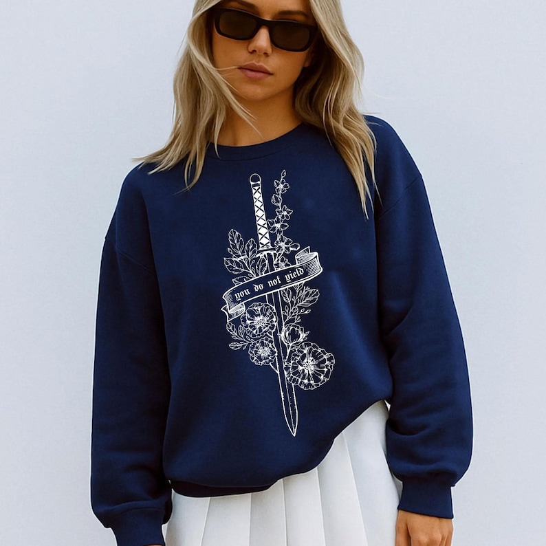 May include: Navy blue sweatshirt with a white graphic of a sword, flowers, and the text "you do not yield." The design is centered on the front of the long-sleeved top. The sweatshirt is a casual style.