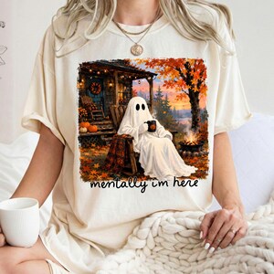 May include: A cream-colored t-shirt featuring a graphic of a ghost sitting in a wooden chair on a porch, holding a mug. The scene includes a campfire, autumn trees with orange leaves, and pumpkins. The text below the graphic reads, mentally i'm here.