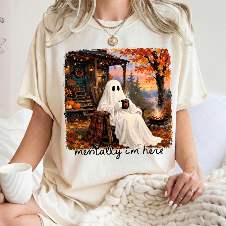 May include: A cream-colored t-shirt featuring a graphic of a ghost sitting in a wooden chair on a porch, holding a mug. The scene includes a campfire, autumn trees with orange leaves, and pumpkins. The text below the graphic reads, mentally i'm here.