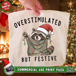 May include: A cream-colored tote bag featuring a cartoon raccoon wearing a Santa hat and tangled in Christmas lights. The text "OVERSTIMULATED BUT FESTIVE" is printed above and below the raccoon illustration. The bag is held up by a person.