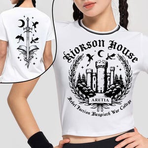 Puede incluir: Top corto blanco con un dise&ntilde;o gr&aacute;fico en blanco y negro. La parte delantera presenta un castillo dentro de una corona, con el texto "Riorkson House" y "Aretia". La parte trasera tiene un dise&ntilde;o de espada, libro y luna creciente.