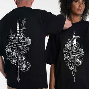 May include: Black t-shirts with a white graphic design. One shirt features a sword with floral accents and the text "you do not yield." The other shirt has a snake wrapped around a sword with floral accents and the same text.