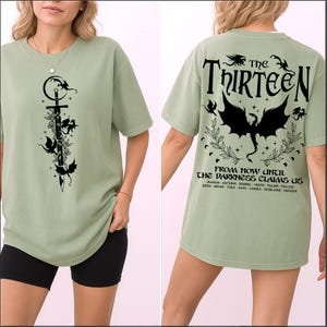 May include: Sage green t-shirt with a black graphic of a sword, dragons, and floral elements on the front. The back features the text "The Thirteen" and "From Now Until The Darkness Claims Us" with a dragon illustration.