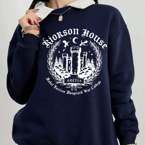 Puede incluir: Sudadera azul marino con cuello blanco y dise&ntilde;o gr&aacute;fico. El dise&ntilde;o presenta el texto "Riorkson House" y "Aretia" con un castillo, &aacute;rboles y una luna creciente. Tambi&eacute;n se incluyen las palabras "Rebel Faction Basgiath War College".