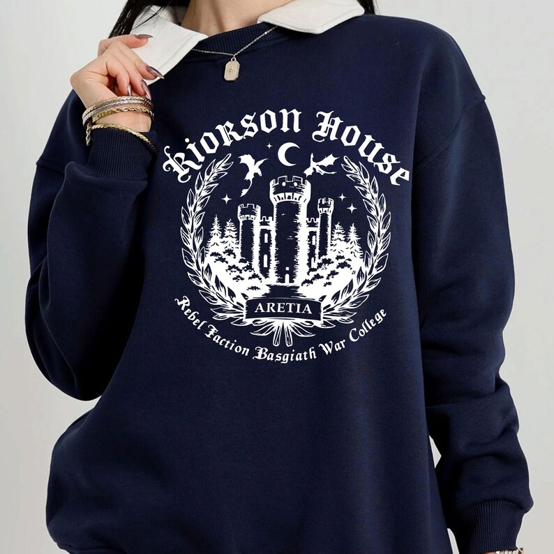 Puede incluir: Sudadera azul marino con cuello blanco y dise&ntilde;o gr&aacute;fico. El dise&ntilde;o presenta el texto "Riorkson House" y "Aretia" con un castillo, &aacute;rboles y una luna creciente. Tambi&eacute;n se incluyen las palabras "Rebel Faction Basgiath War College".