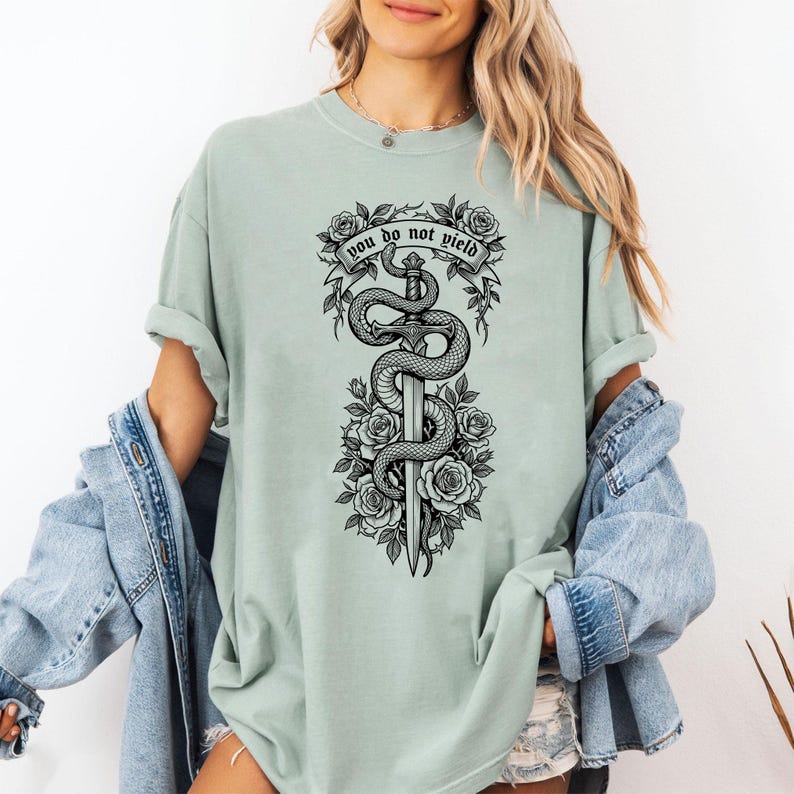 May include: A light green t-shirt featuring a black and white graphic of a sword entwined by a snake, surrounded by roses. The text "you do not yield" is above the sword. The shirt has short sleeves and a relaxed fit.
