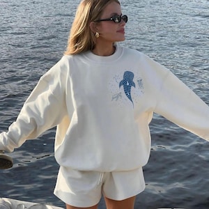 May include: Cream-colored sweatshirt and shorts set. The sweatshirt features a blue whale shark graphic with other sea creatures. The shorts have a drawstring waist. The outfit is worn by a person with sunglasses.