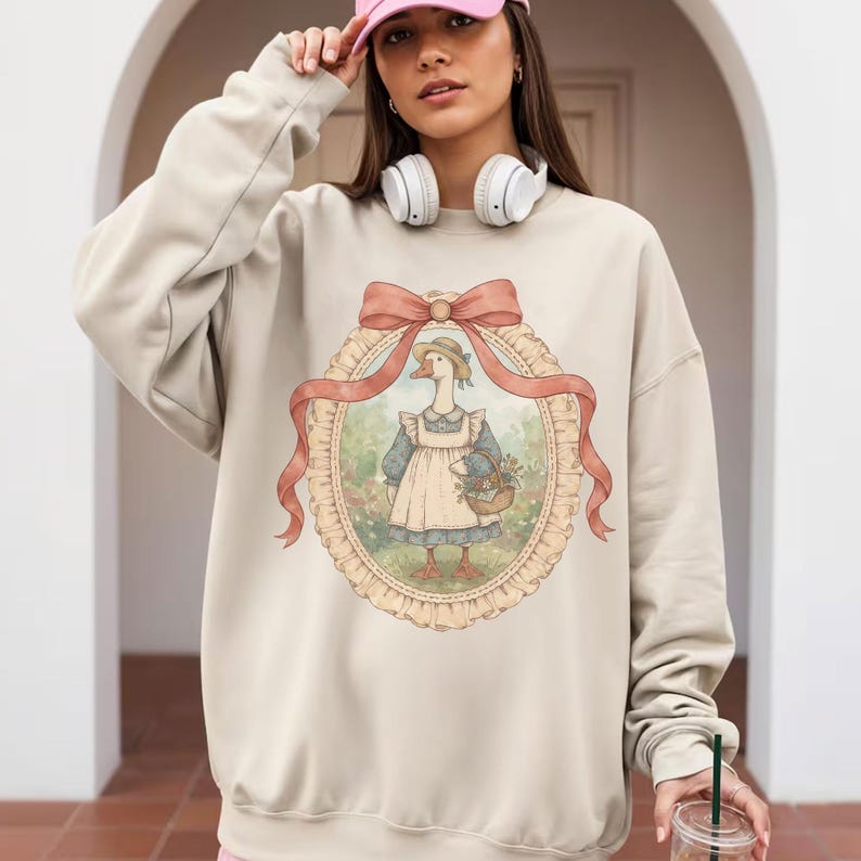 May include: Beige sweatshirt featuring a framed illustration of a goose wearing a bonnet and apron, holding a basket of flowers. The illustration is surrounded by a pink ribbon and bow. The sweatshirt has a crew neck and long sleeves.