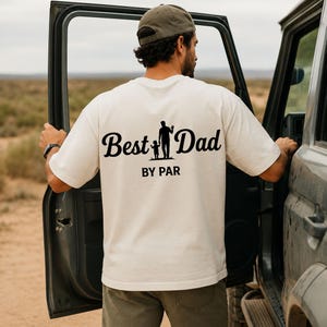 May include: Cream-colored t-shirt with the words "Best Dad" in a cursive font, accompanied by a silhouette of a father and child, and the text "BY PAR" below. The shirt is worn by a person standing next to a vehicle.