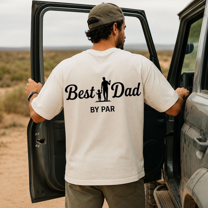 May include: Cream-colored t-shirt with the words "Best Dad" in a cursive font, accompanied by a silhouette of a father and child, and the text "BY PAR" below. The shirt is worn by a person standing next to a vehicle.