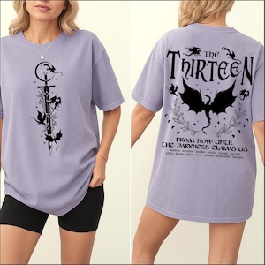 May include: A lilac-colored t-shirt with a black graphic on the front featuring a sword, flowers, and winged creatures. The back of the shirt displays the text "THE THIRTEEN" and "FROM NOW UNTIL THE DARKNESS CLAIMS US."