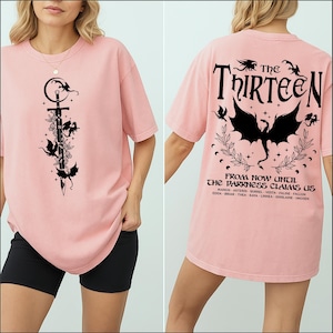 May include: A light pink t-shirt featuring a black graphic of a sword, flowers, and bats. The back of the shirt displays the text "The Thirteen" above a bat graphic and the phrase "From Now Until The Darkness Claims Us."