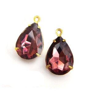 May include: Two teardrop-shaped crystal pendants with gold-tone settings. The crystals are a deep purple color and have a faceted design.