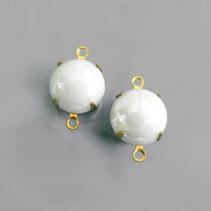 May include: Two white, round cabochon beads with a pearlescent sheen. Each bead is set in a gold-coloured metal setting with four prongs and a loop at the top and bottom, suitable for jewellery making.