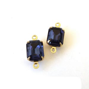 May include: Two rectangular, dark blue crystal pendants with gold-colored metal settings. Each pendant has a loop at the top and bottom for attaching to jewelry. The crystals have a faceted appearance.