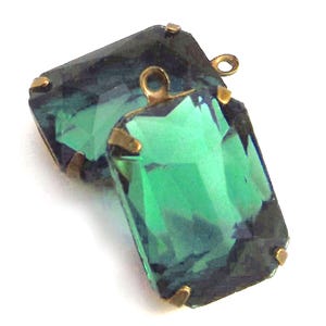 May include: Two emerald-green rectangular crystal pendants with gold-colored metal settings. Each pendant has a loop at the top for attaching to a chain or other jewelry component. The crystals have a faceted cut, reflecting light.