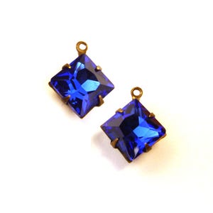May include: Two square, faceted, royal blue crystal pendants with brass-colored metal settings. Each pendant has a small loop at the top for attaching to a chain or other jewelry component. The crystals are a vibrant, saturated blue.