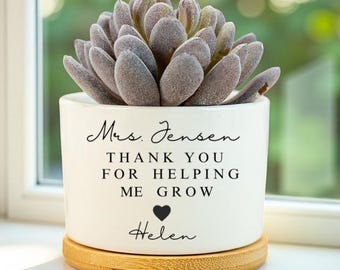 May include: A white ceramic planter with a succulent plant. The planter has the text "Mrs. Jensen, Thank You For Helping Me Grow" with a heart and the name "Helen". The planter sits on a round, wooden base.