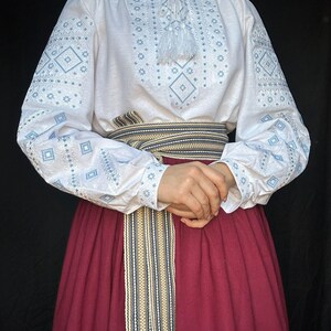 Puede incluir: Una blusa blanca bordada con motivos geom&eacute;tricos azules y una borla blanca. La blusa se lleva con una falda color burdeos y un cintur&oacute;n tejido con rayas beige, azules y negras.